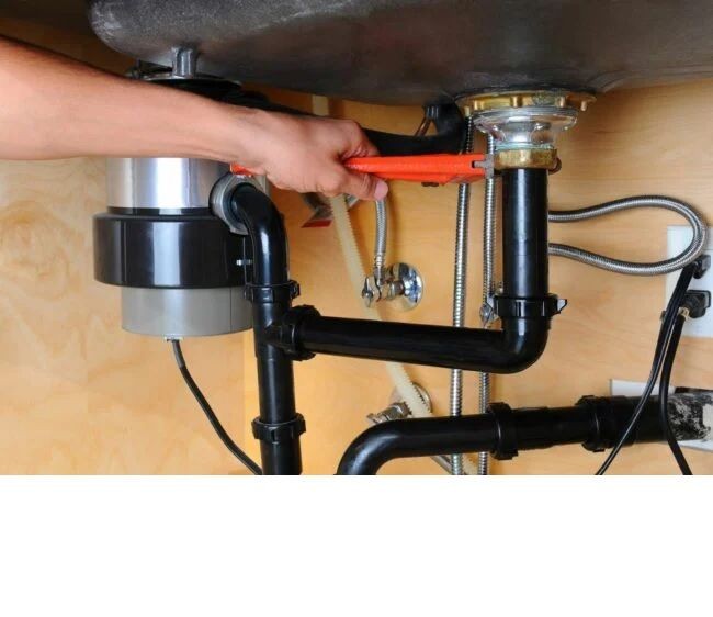 Hand using a wrench to tighten pipe fittings under a kitchen sink with plumbing and disposal unit visible.