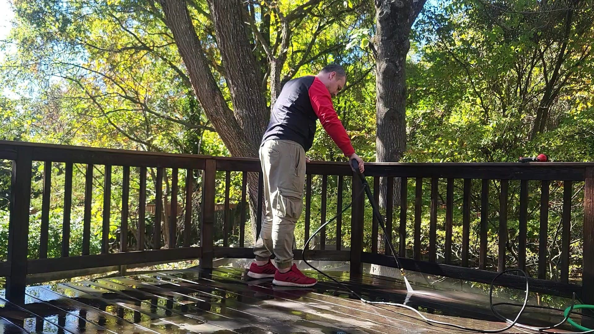 Person using a pressure washer to clean a wooden deck surrounded by trees on a sunny day.